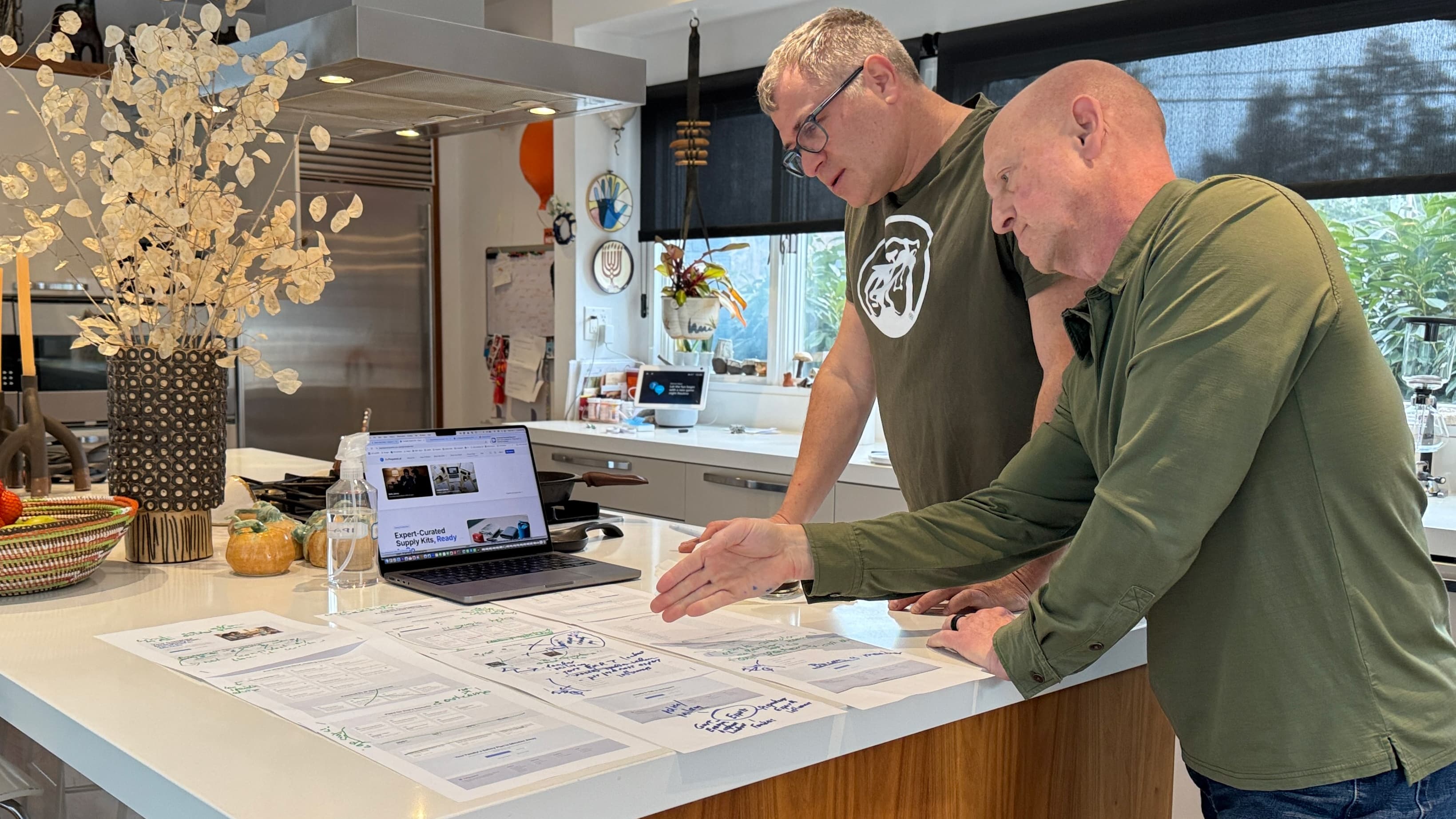 Brian and Tiran reviewing the BePreparedSolutions.co website at a kitchen island, with printed wireframes and hand-written annotations spread across the counter