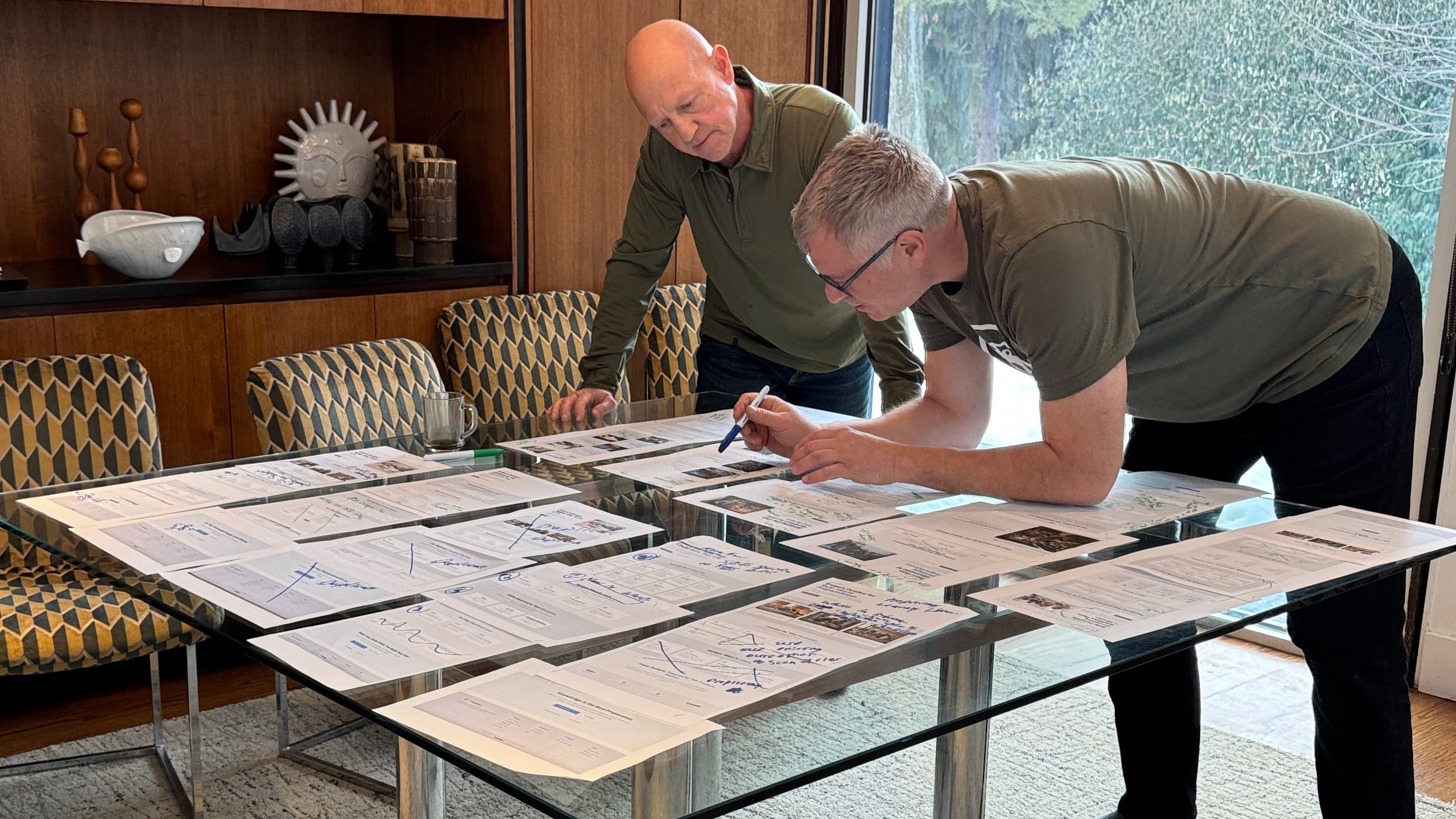 Brian and Tiran working over a glass dining table covered in annotated printouts with crossed-out sections and hand-drawn diagrams